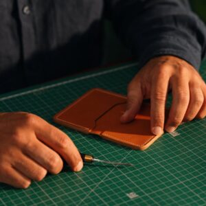 Close-up of hands crafting a leather wallet on a green cutting mat, emphasizing artisanal craftsmanship.