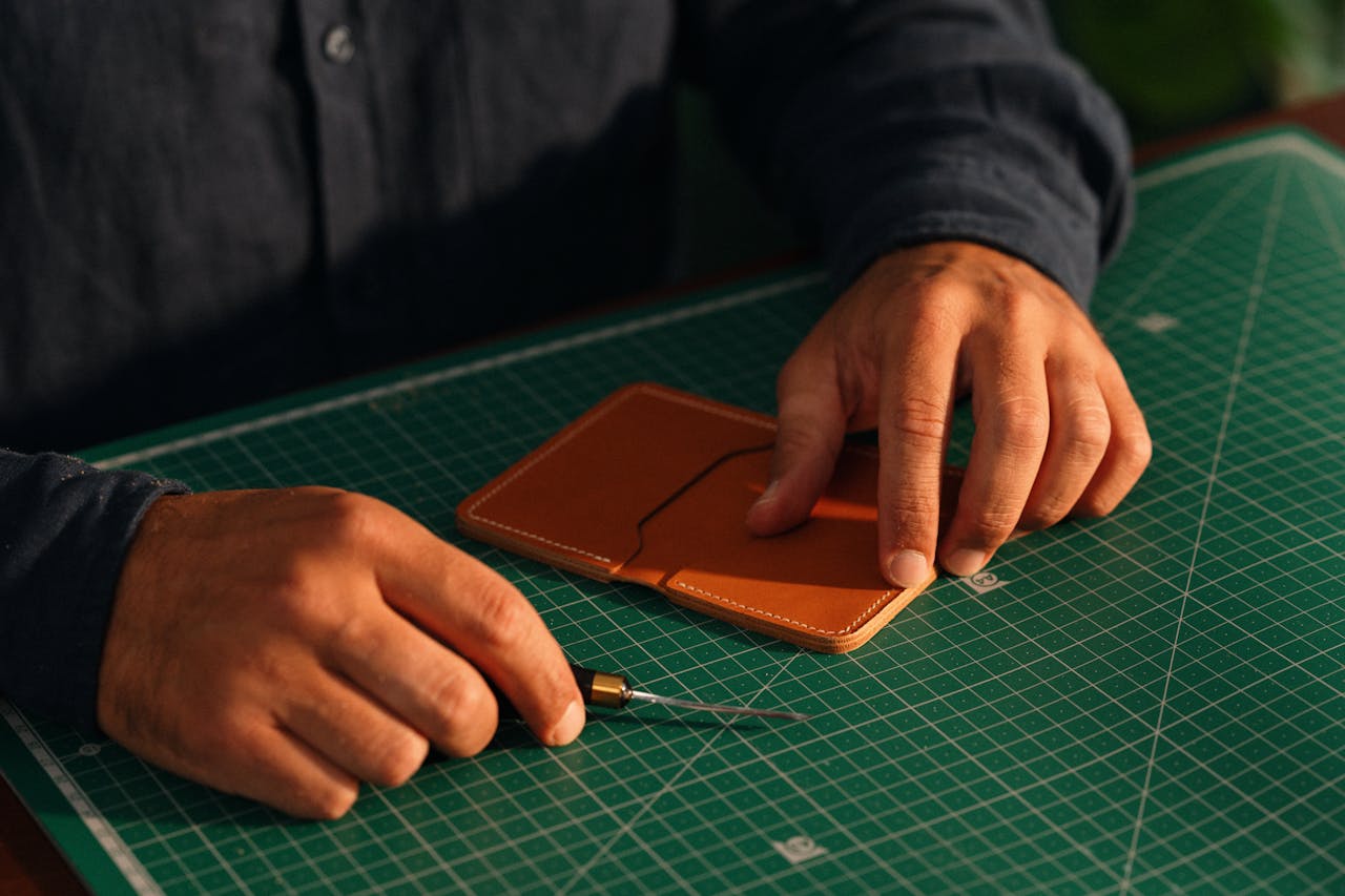 services-04 Close-up of hands crafting a leather wallet on a green cutting mat, emphasizing artisanal craftsmanship.